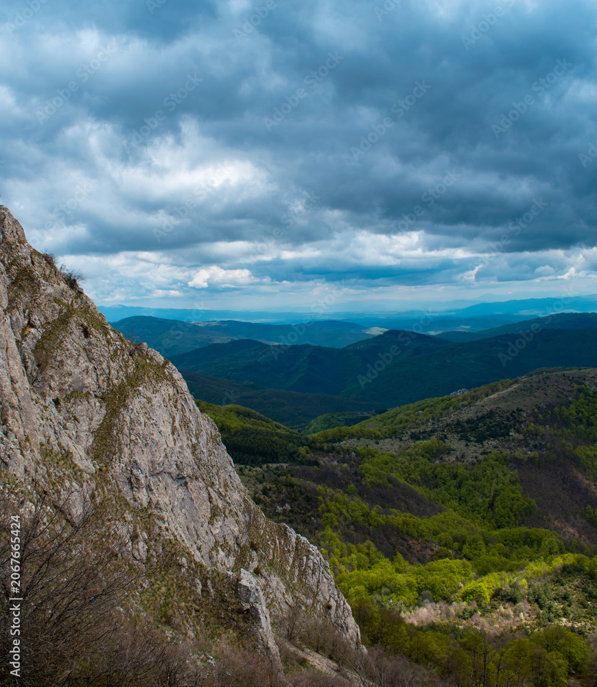 Fototapeta premium Dramatic mountain view from a rocky cliff overlooking green valleys under a stormy, cloudy sky. Epic landscape of high green mountains and rocky peak before a storm.