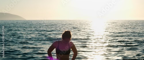 Woman enjoying sunset on the beach, standing calmly with golden backlight and gentle sea waves, anamorphic static shot.
