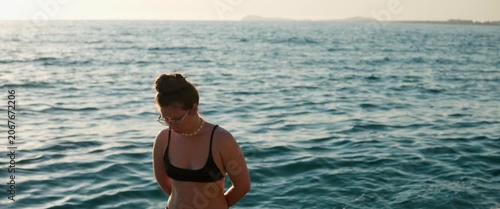 Static anamorphic medium shot of woman in bikini standing by the sea and sunbathing from a different angle. Calm water, soft evening light and relaxed pose create a peaceful summer beach mood.