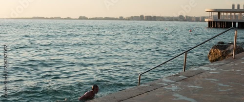 Man walking up stone steps out of the sea at sunset, wide static anamorphic shot with city skyline and pier in background.