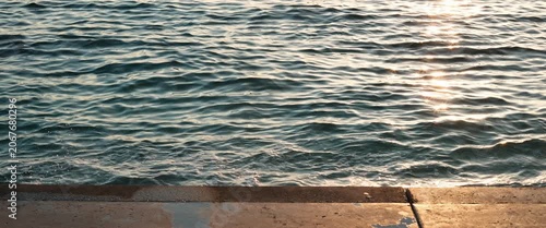 Calm sea waves rippling at sunset with golden sunlight reflecting on the water near a stone pier, static anamorphic shot.