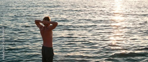 Static anamorphic medium shot of shirtless man standing by sea at sunset and sunbathing. Back view, hands behind head and shimmering water create a calm, peaceful and serene coastal atmosphere.
