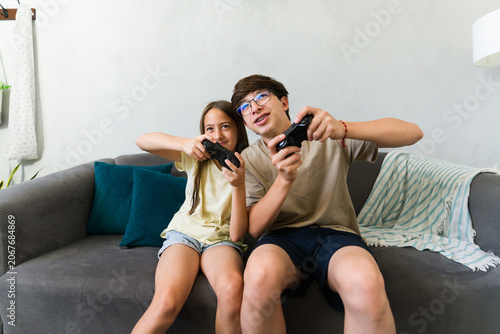 Siblings playing video games at home, brother and sister enjoying fun leisure time together on the sofa