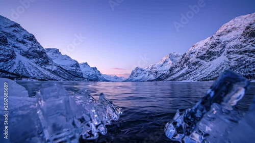 Icy crystals glisten on the water's edge as majestic mountains rise in the background. Soft dawn light casts serene shades of blue, inviting stillness and awe in this breathtaking landscape.