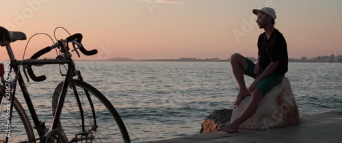 Static anamorphic wide shot of man in T shirt sitting on a stone by the sea and watching the sunset. Bicycle in foreground and calm water emphasize a thoughtful, peaceful coastal leisure mood.