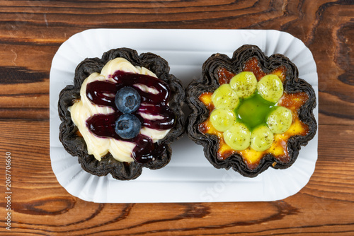 Two flower-shaped black crust egg tarts topped with blueberry cream sauce and green grape fruit glaze on white paper tray