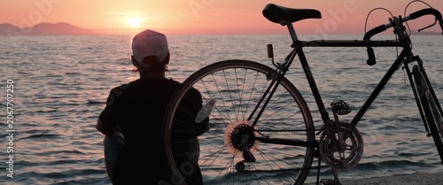 Vintage bicycle in close-up foreground as a man sits down to watch the sunset over the sea, anamorphic static shot.