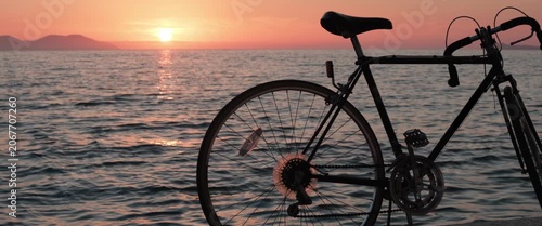 Static anamorphic wide shot of bicycle silhouette parked by the sea at sunset. Orange sky, ocean horizon and glowing reflections create a calm coastal travel and leisure atmosphere.
