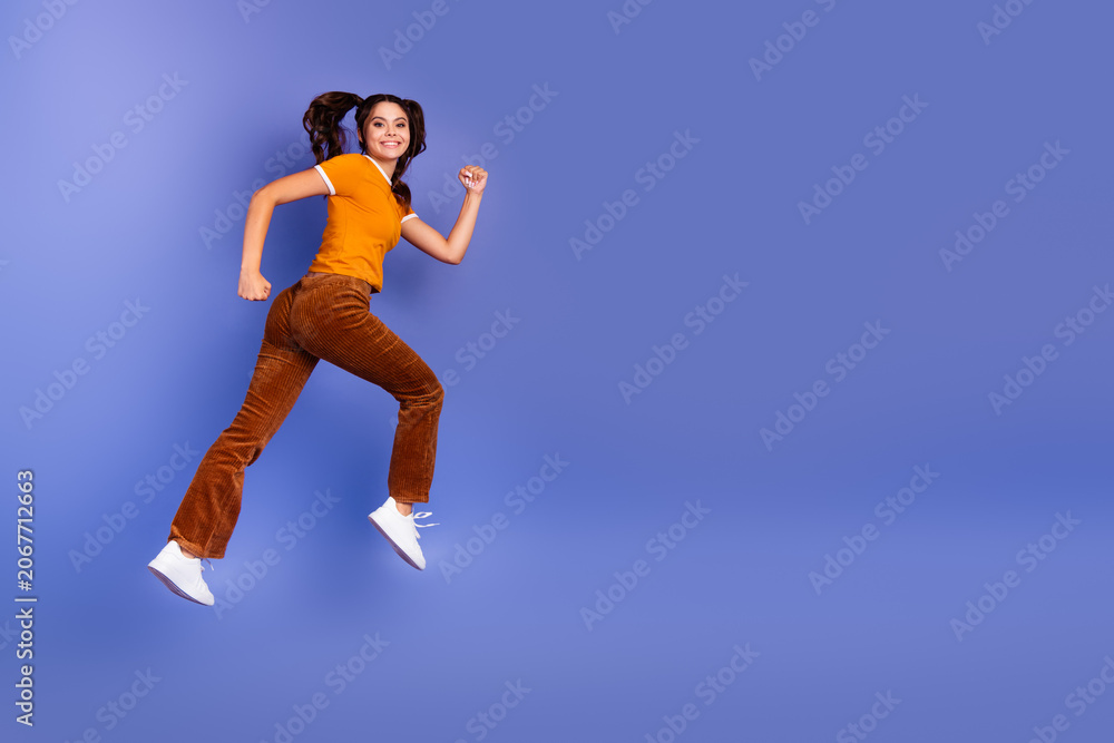 Naklejka premium Jumping young girl in orange shirt and brown pants against blue background expressing happiness and style
