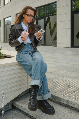 Foto Young woman in leather jacket sitting on urban bench checking her phone with a r