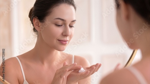 Young woman applying moisturizing cream to her hand while looking in mirror. Daily skincare routine and dermatological care concept.
