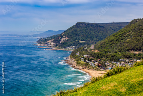 A beautiful aerial view of Clifton and Sea Cliff Bridge with sandy beach and waves crashing on the shore. The beach is bordered by lush green cliffs and the bright blue sky.