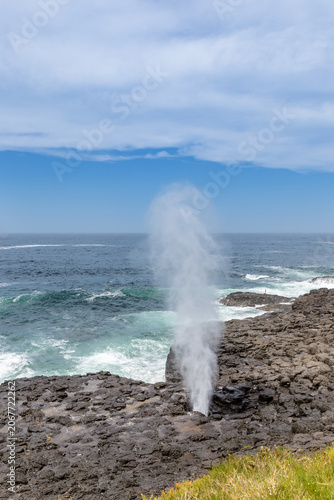 A powerful jet of water erupts from the Little Blowhole in Kiama, New South Wales, Australia, against a backdrop of blue sky and crashing waves.
