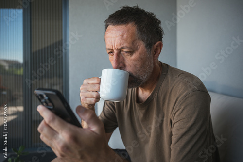 Middle-aged man drinking coffee while checking smartphone on balcony at home in morning light