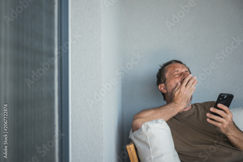 Tired middle-aged man sits on a balcony sofa, yawning and covering his mouth while checking his smartphone, conveying fatigue, boredom, late-night scrolling and everyday digital life.