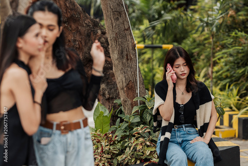 Фотография A young Southeast Asian woman sits alone talking on her phone unaware that two women talk behind her back and slander her