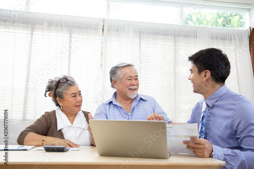 A cheerful elderly Asian couple sits at a desk, smiling while consulting a professional financial advisor or insurance agent holding documents in front of a laptop computer at home