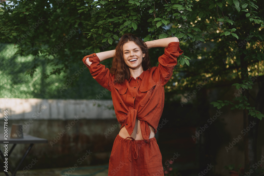 Fototapeta premium Young woman with curly brown hair laughs with arms raised in a rust colored matching set outdoors, enjoying a carefree moment in dappled shade, sunlight warming her bright smile on a summer day