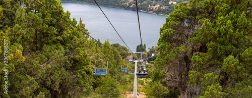 Schilderij op canvas Panoramic view of chairlift moving down a mountain forest with a deep blue lake