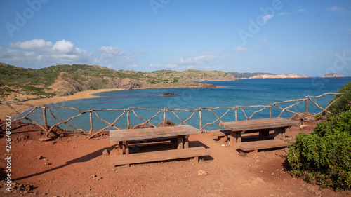 Two wooden picnic tables overlooking a scenic bay with a sandy beach and hills