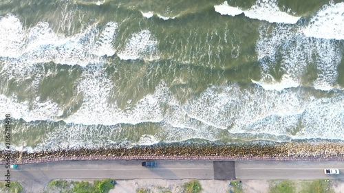 Wave struck seawall Surfside Beach frontage along Seashell Dr shows strong surf hitting rock lined barrier beside Gulf facing roadway. Sunlit water, bright foam highlights extend, Freeport, Texas