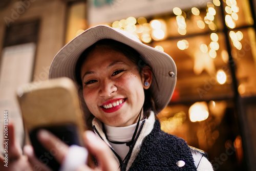Authentic portrait of smiling Asian woman holding smartphone, Chinese traveler looking away in evening city street, 5G mobile connectivity, authentic person lifestyle, copy space.