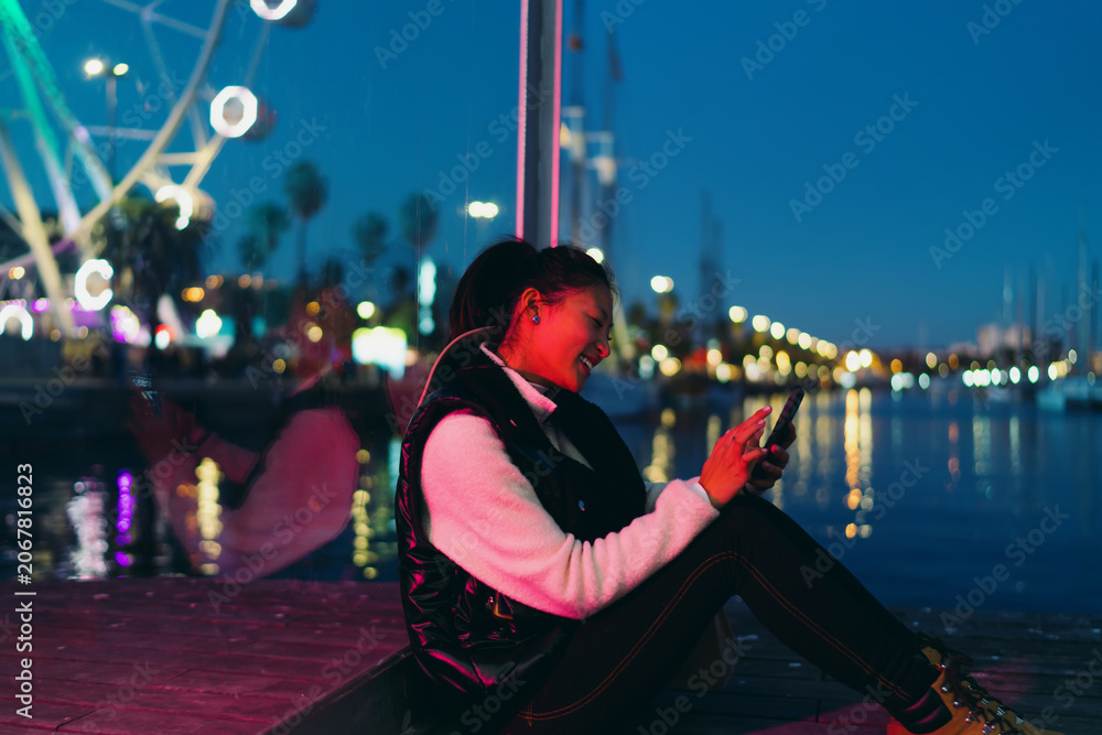Obraz premium Asian woman using smartphone application on night city pier with neon lights. Smiling female tourist browsing social media on mobile phone with bokeh ferris wheel background, digital nomad lifestyle.