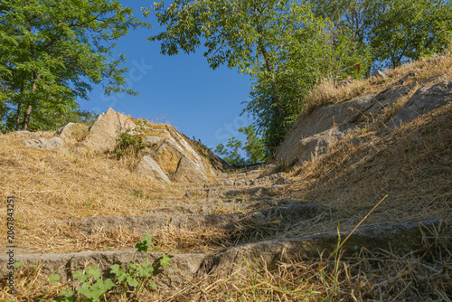 Stone steps climbing a dry hillside between rocks and trees under clear blue summer sky