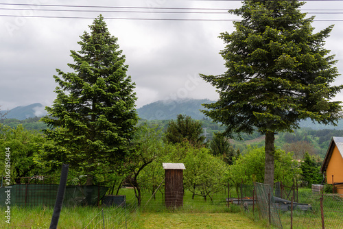 Traditional wooden outhouse stands among fruit trees and garden plots beneath misty mountains and Norway spruce trees. The rural landscape combines village gardening, backyard structures, orchard vege