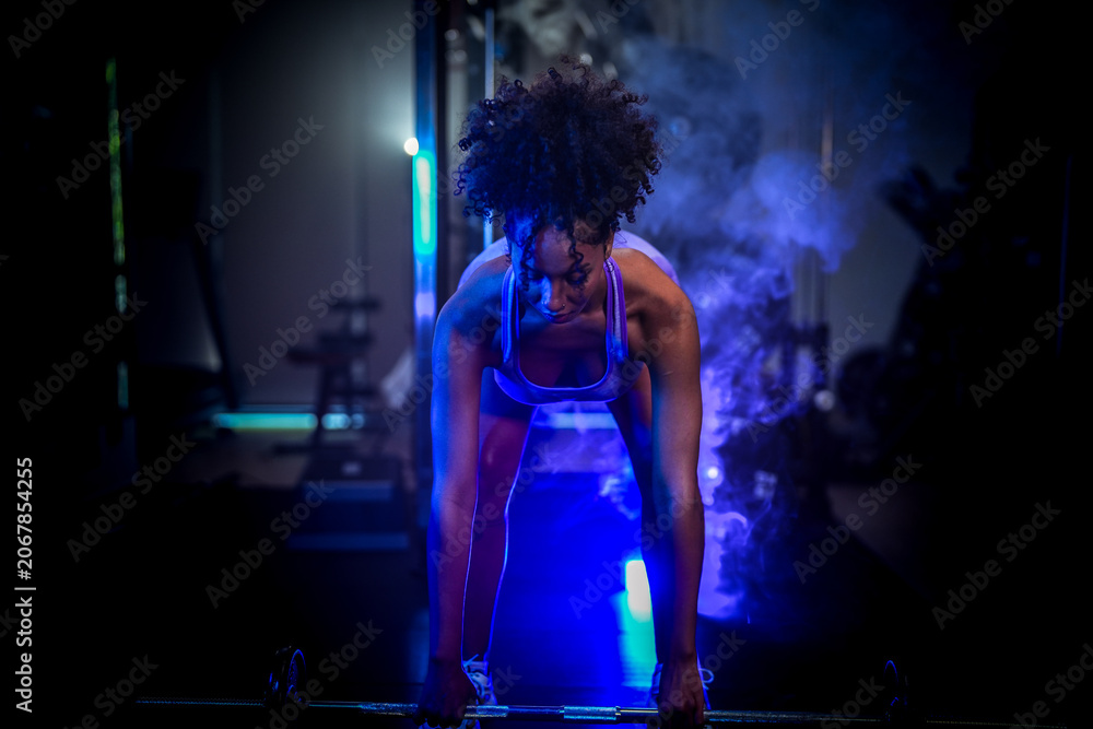 Naklejka premium A woman in purple sportswear preparing for a heavy lift with a barbell in a moody, low-light gym environment with dramatic highlights.