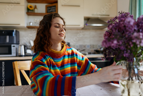 Foto A woman sits at a sunny kitchen table, reaching toward a bouquet of flowers