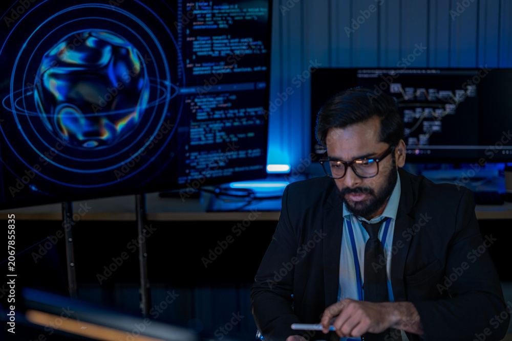 Fototapeta premium South Asian male developer in a white shirt uses a tablet device while surrounded by screens displaying algorithms in a dark tech office.