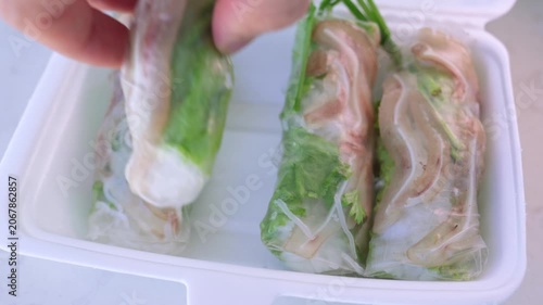 Hand taking a fresh vietnamese spring roll, filled with pork ear, vegetables, and rice noodles, from a white disposable container, presenting a popular and healthy asian appetizer