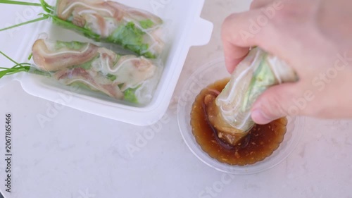 Human hand dipping a traditional vietnamese spring roll, filled with pork ear and fresh vegetables wrapped in rice paper, into a savory peanut dipping sauce from a takeout container