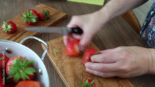 Slicing fresh organic strawberries on olive wood cutting board in kitchen. Hands of chef cutting summer berries with knife next to enamel colander reflecting culinary creativity and food styling