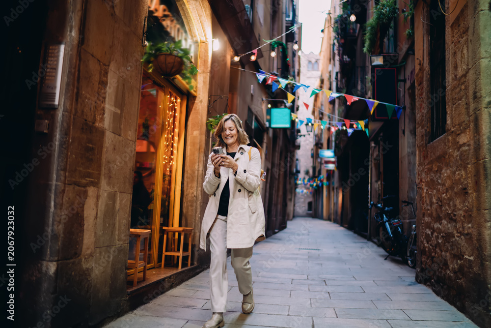 Obraz premium Full length smiling woman using smartphone walking on street. Mature traveler checking booking, mobile banking, online service, digital nomad lifestyle, authentic representation, copy space