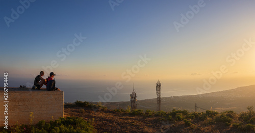 Mother and son sitting on concrete platform at sunrise, looking at scenic panoramic view towards Malia town from Mount Edheri in Crete, Greece. Family hiking and travel concept