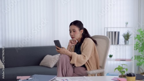 Young Asian woman looking at smartphone with concerned expression while working remotely in bright modern home office