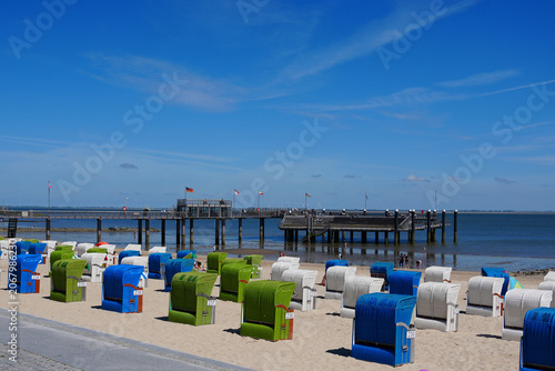 A lively coastal scene in Wyk auf Föhr, Germany. Tourists and locals relaxing on the sandy beach and promenade next to traditional strandkorb chairs and the famous Mittelbrücke pier at the North Sea.