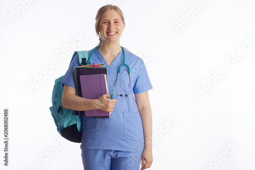 A young woman in medical scrubs carries a stack of notebooks and a pen, representing the academic side of nursing school, exam preparation, and clinical studies.
