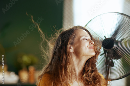 A smiling woman with closed eyes and flowing brown hair tilts her head back, enjoying a refreshing breeze from an electric fan. This indoor scene captures deep relief and well-being from hot weather.