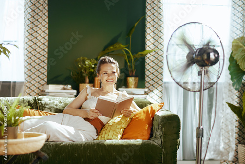 A smiling woman with a book finds peace and cool comfort on a green velvet sofa in her sunlit living room, enjoying the refreshing breeze from a standing fan on a warm day.