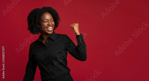 Excited young black woman celebrating success with raised fist showing winning gesture over isolated red background with copy space