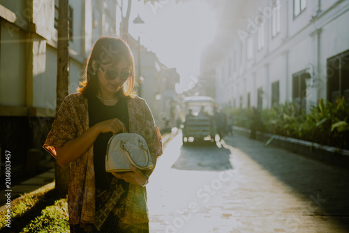 Fashionable adult Asian woman in sunglasses and layered outfit with batik bottom opening a small her bag standing on alley city street during sunsets.