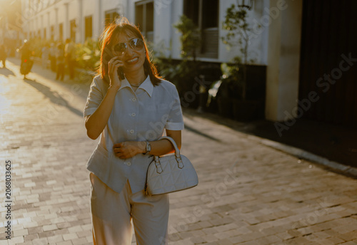 Fashionable style adult Asian Businesswoman in sunglasses and casual button down shirt talking by phone call while walking in city at sunsets.