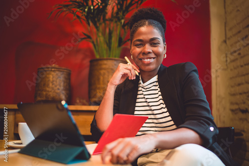 Smiling Black woman holds stylus and red notebook at cozy café table with tablet, representing confident freelance work, digital creativity, and joyful focus in modern business setting.