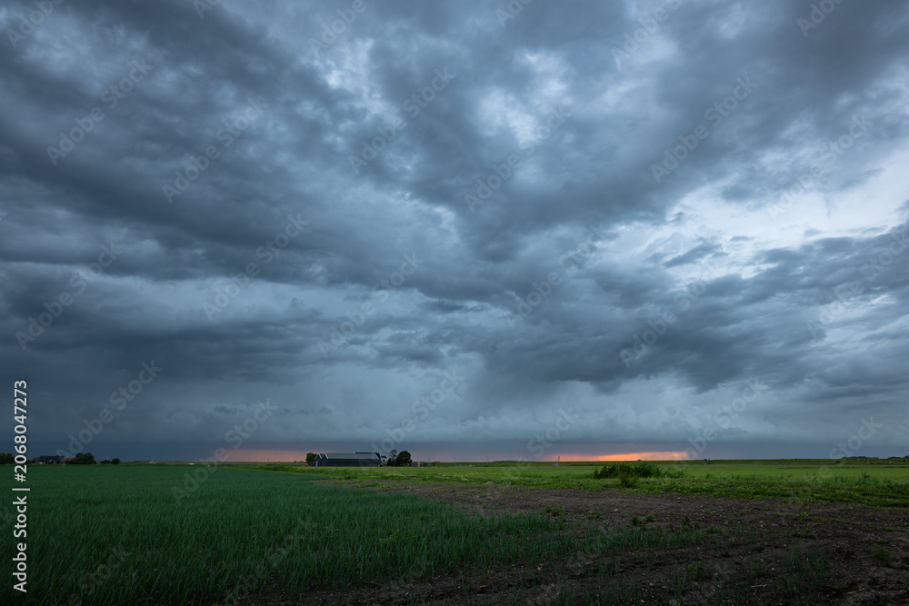 Fototapeta premium Dramatic dark storm clouds sweep over a flat rural farmland as a thin band of sunset light glows on the distant horizon