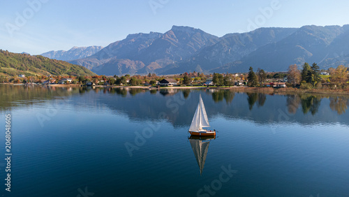 Aerial 4K drone view of a sailboat on calm Lake Caldonazzo, Italy. Features perfect mirror reflections of the scenic Trentino mountains and autumnal shoreline on still blue water.