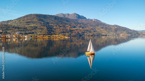 Aerial 4K drone view of a sailboat on calm Lake Caldonazzo, Italy. Features perfect mirror reflections of the scenic Trentino mountains and autumnal shoreline on still blue water.