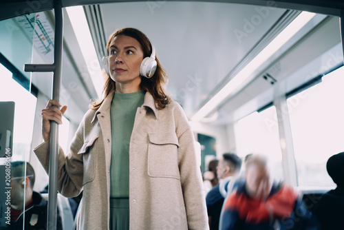 Middle aged Caucasian woman standing in moving tram, wearing wireless headphones, looking out window thoughtfully, public transport commuting, urban lifestyle, authentic passenger portrait.